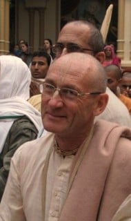 Portrait of an older person wearing glasses and a light shawl in a crowded courtyard, smiling among peers; image accompanies an article on book distribution, community outreach, and lifelong dedication.