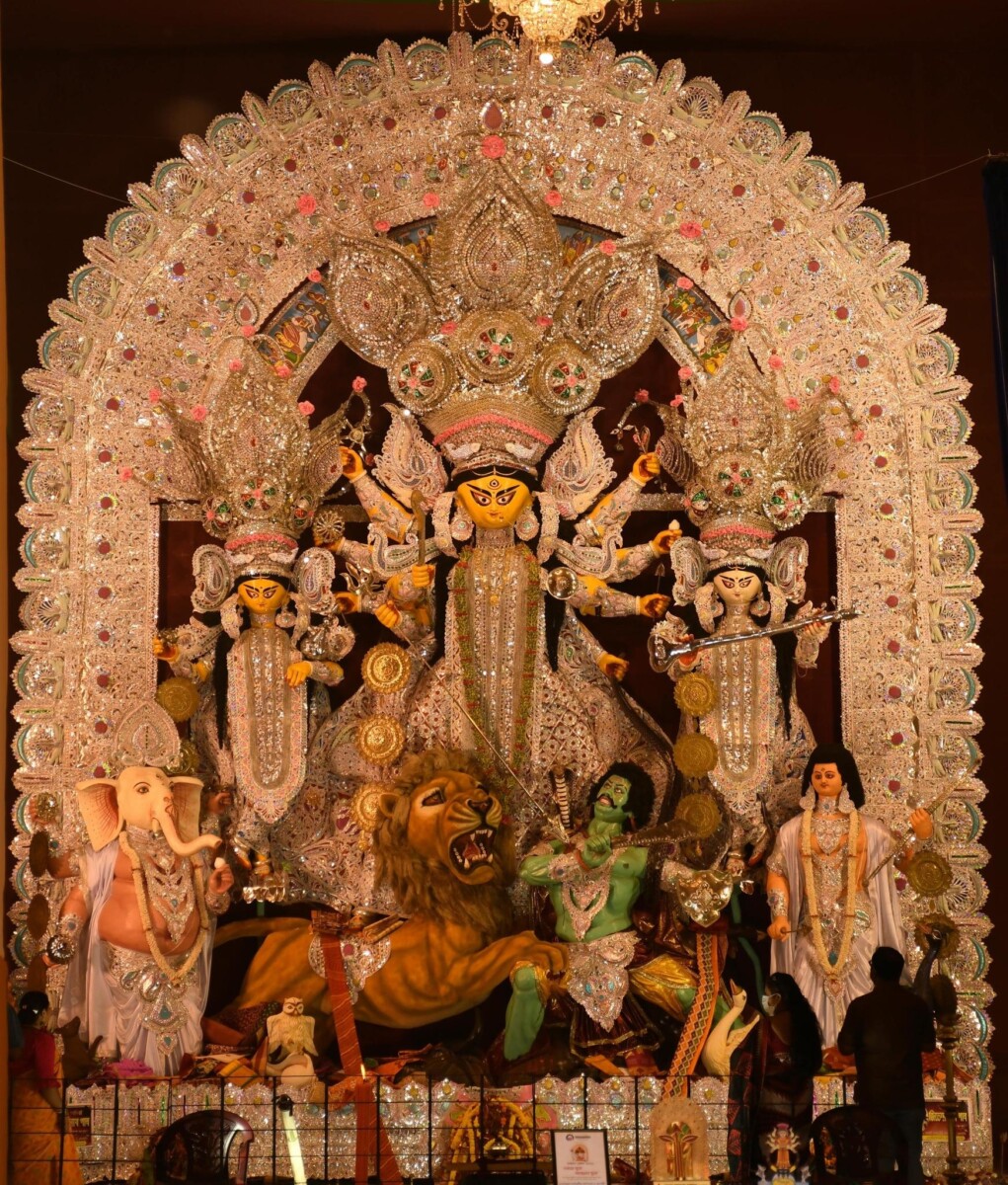 Durga Puja idol tableau: Durga on a lion slays Mahishasura, flanked by Lakshmi, Saraswati, Ganesha and Kartikeya beneath an ornate arch; site of the Kola Bou (Nabapatrika) ritual honoring Prakriti and Shakti.