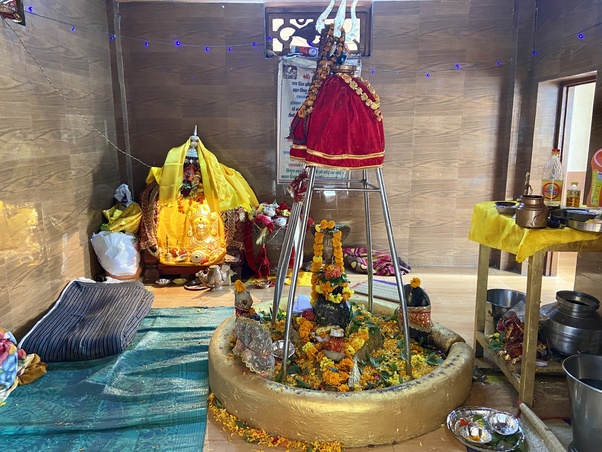 Interior of a Himalayan temple in Uttarkashi: a marigold-covered Shiva lingam under a metal canopy, nearby idols in red and yellow cloth, floor mats, and brass offering vessels.