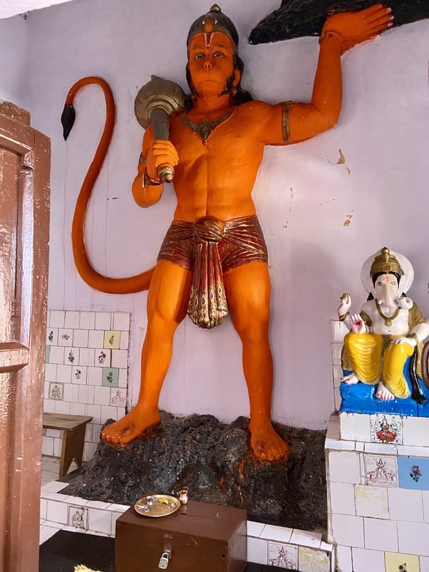 Interior of a Himalayan shrine: a tall orange Hanuman statue holds a mace and raises one hand, tail curled behind. A smaller white-and-gold Ganesha sits on a tiled altar; offerings and a donation box below.