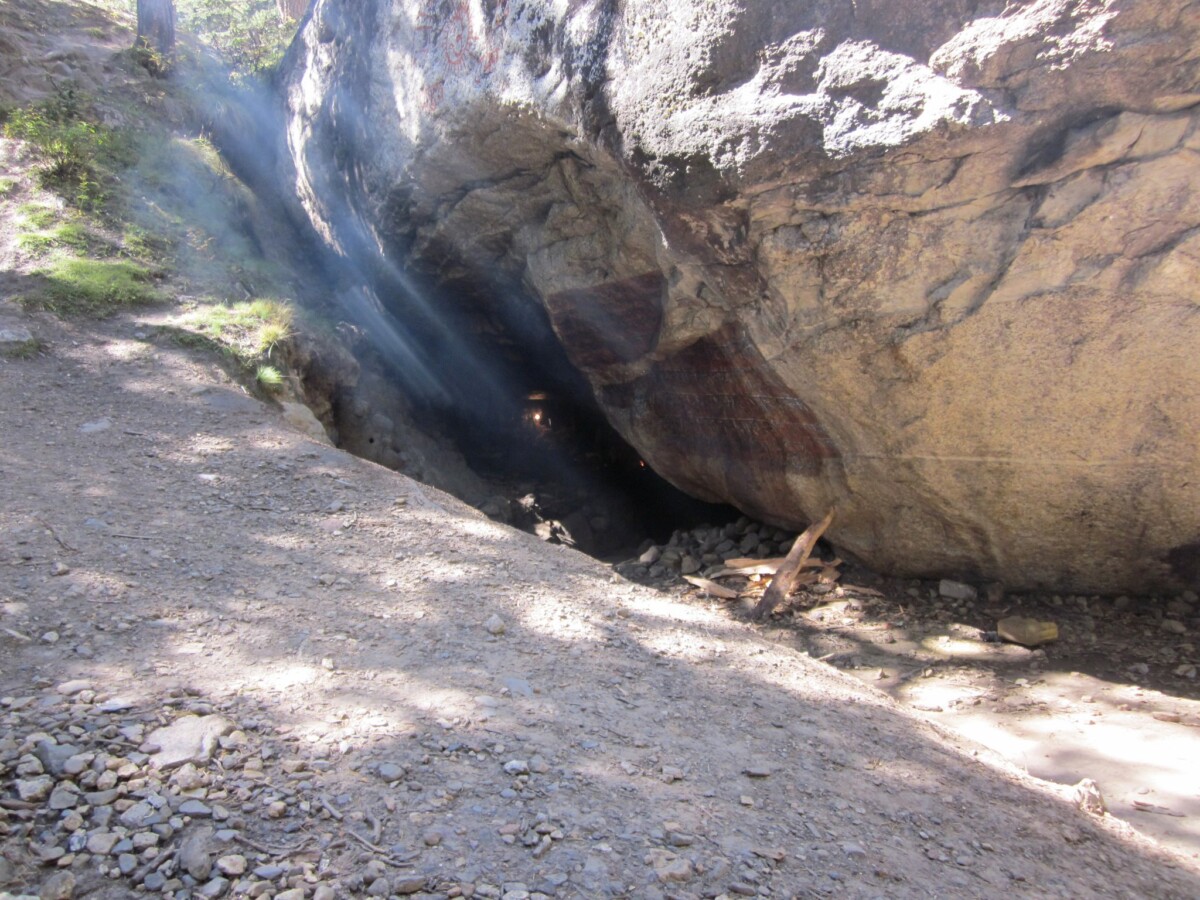 Sunlight slants into a small cave beneath a massive boulder beside a rocky footpath on the Gangotri–Gaumukh trek in the Himalaya, revealing rugged textures and quiet stillness.