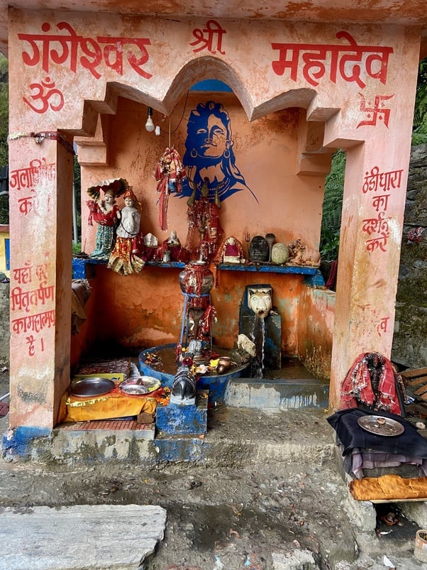Small roadside shrine in Gangnani village with a Shiva lingam under a metal naga hood, Mahadev mural, Hindi lettering, offerings, and a cow-head water spout in the Himalayan foothills; Living Dharma travel.