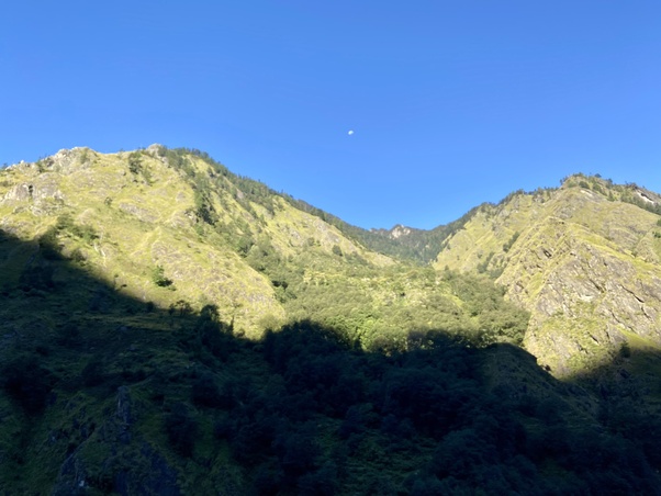 Sunlit ridges above Gangnani in the Hindu Himalaya, with forested slopes and rocky outcrops under a cloudless blue sky; a faint moon hangs overhead while the valley foreground lies in deep shadow. Living Dharma travel.