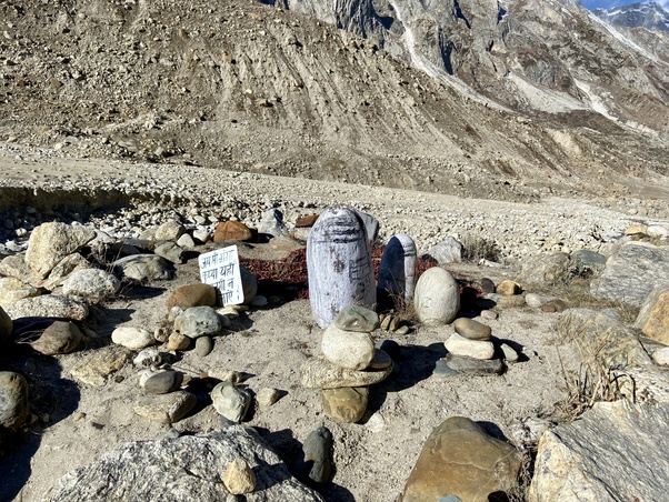 Stone Shiva lingam shrine with cairns and a small Hindi sign on the rocky riverbed near the Gangotri–Gaumukh trail in the high Himalaya, framed by barren slopes and glacier-scarred mountains.