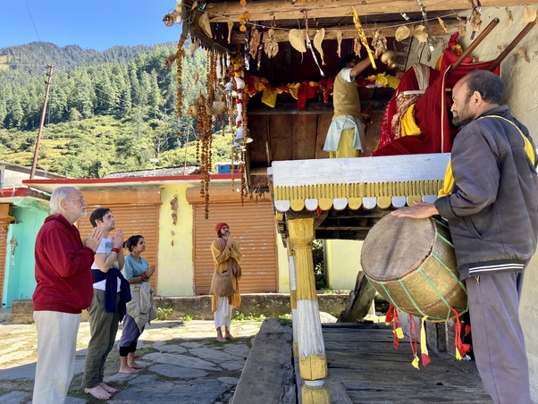 Devotees stand barefoot before a small Himalayan temple in Gangnani Village as a drummer plays and a priest performs ritual offerings beneath garlands and bright cloth, with pine-covered mountains behind.