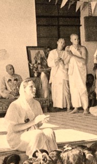 Sepia-toned photo of a kirtan gathering in a hall: a seated lead singer gestures mid-chant while a crowd listens; robed devotees stand behind near a framed portrait and bunting, evoking historic devotional music in India.