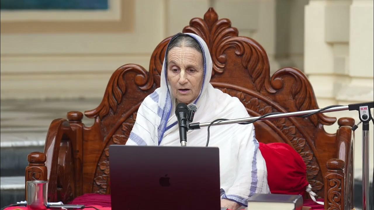 Devotee in a white sari speaks into a microphone from an ornate wooden seat, using a laptop and scripture, delivering a temple lecture on the Disappearance Day of Sri Vasudeva Ghosh (SB 10.85.41-13) for our testing series.