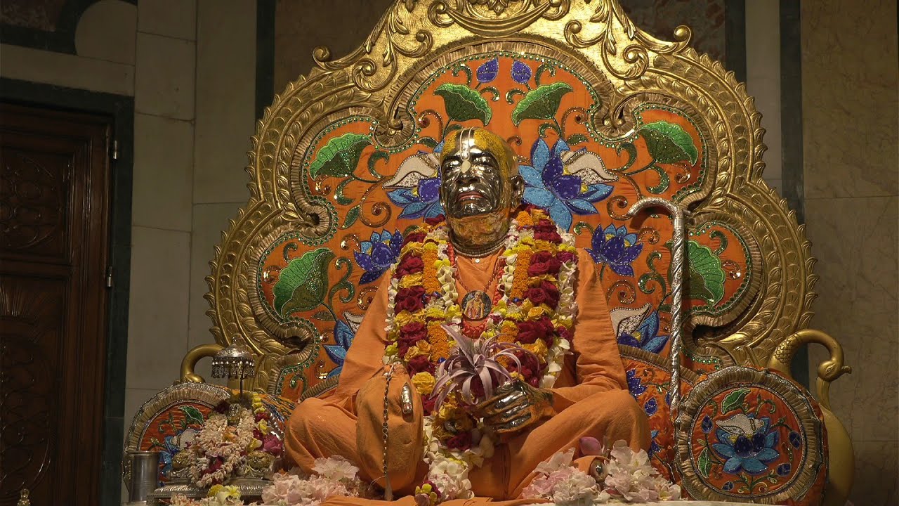 Garlanded statue of a revered spiritual teacher seated cross‑legged on a golden throne with lotus and leaf motifs, surrounded by flowers, offerings, and a silver staff during a devotional ceremony, testing.