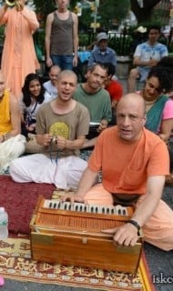 Park kirtan gathering with a harmonium on a rug as participants sit nearby, chanting, clapping, and counting mantra beads—an authentic moment highlighting transcendental sound vibration in practice.