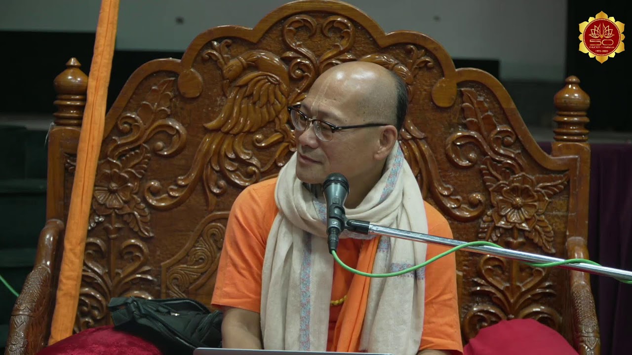 Spiritual teacher in saffron attire seated on an intricately carved wooden throne, speaking into a microphone during a C.C. Adi Lila 4.170 class at ISKCON Vrindavan; photo for testing.