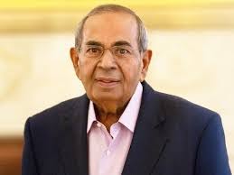 Portrait of an elderly man with glasses, wearing a navy blazer and light pink shirt, facing the camera against a softly lit indoor background; for a memorial update in ISKCON-related testing news.