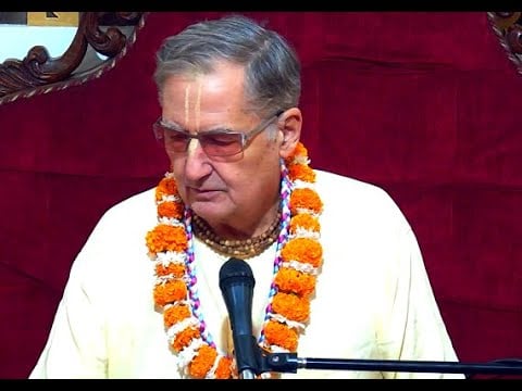 Vaishnava speaker in cream robes with tilak, tulsi beads, and marigold garlands addresses a mic before a red backdrop during a Srimad-Bhagavatam 12.13.13 class at ISKCON Dwarka; testing post.