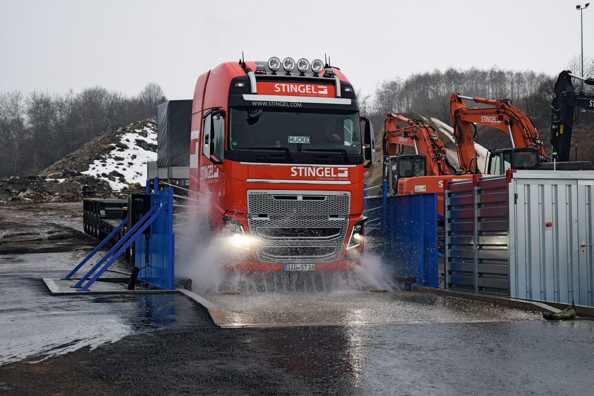 Red heavy-duty truck drives through an automated wheel-wash at a construction site, water spraying around its tires to remove mud and dust; excavators and snowy soil piles line the wet industrial yard.