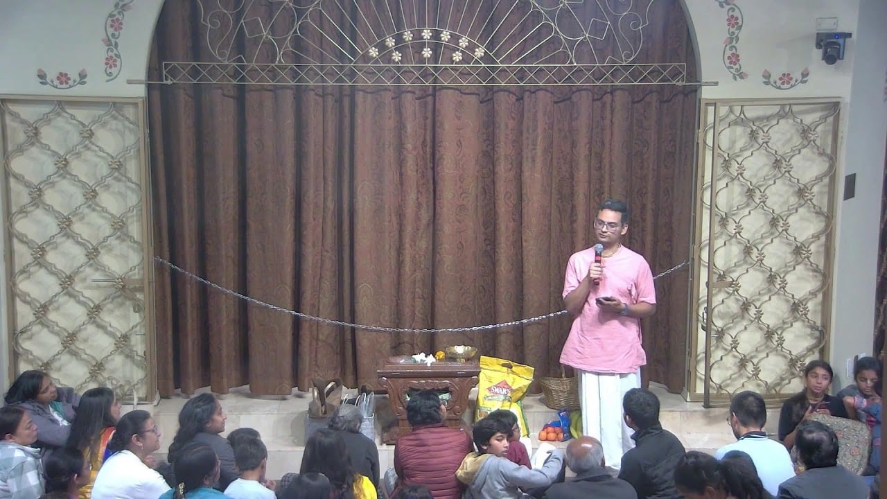In an ISV temple hall, a speaker in pink kurta and white dhoti addresses a seated crowd before ornate curtains, holding a microphone beside fruit and flower offerings during a Krishna Katha. testing