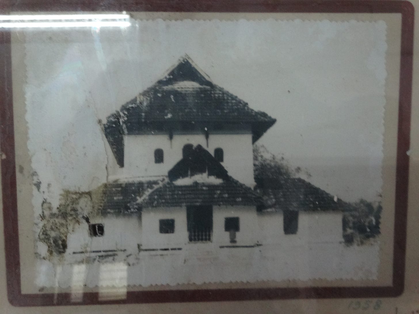 Archival black and white photo of a Kerala style mosque with tiered tiled roofs, small arched windows, and a central gable in a worn frame, evoking the Cheraman Perumal legend and early Islam in Kerala.