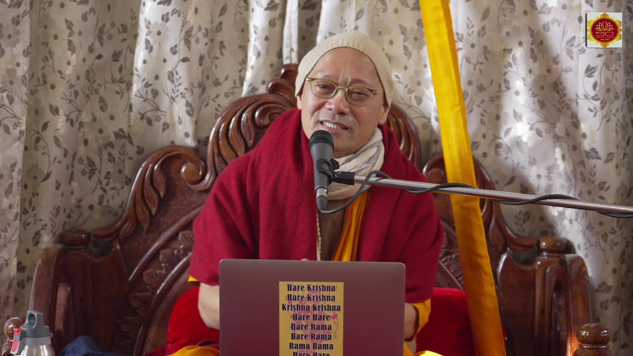 A saffron-robed monk in a red shawl speaks into a microphone from a carved wooden seat; a laptop with the Hare Krishna mantra sits before him, floral curtains behind, during a talk at ISKCON Vrindavan.