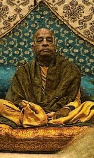 Elderly person seated cross-legged on cushioned dais, eyes closed in meditation, wrapped in ochre robes before a patterned fabric backdrop; contemplative scene for Articles on society, friendship and love.