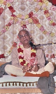 An elderly monk sits cross-legged on a cushioned dais, draped in flower garlands before a richly floral backdrop, speaking into a microphone; image for an article on Uddhava's shock and joy.