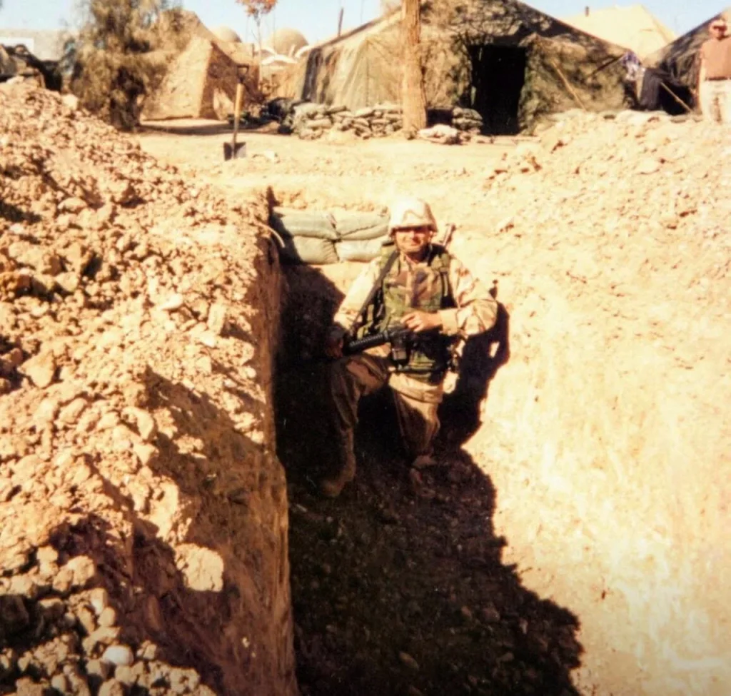 Soldier in camouflage crouches with a rifle inside a narrow desert trench near sandbags and canvas tents, harsh midday light casting sharp shadows across dry, rocky earth.