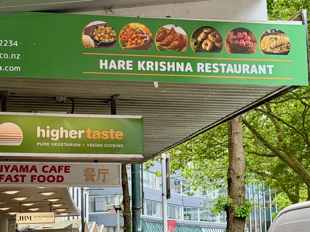 Street view in Wellington of the Higher Taste restaurant sign for Pure Vegetarian & Vegan Cuisine, beneath a larger Hare Krishna Restaurant banner showing dish photos and green branding.