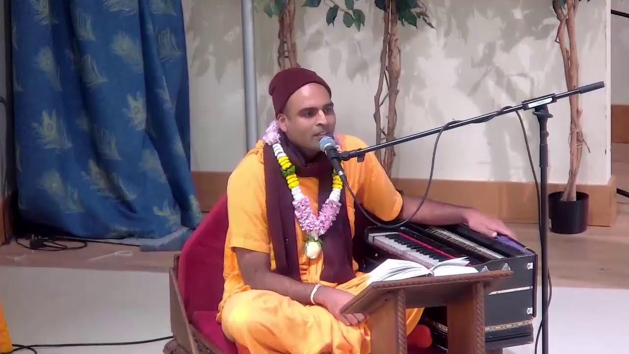 A seated speaker in saffron robes, wearing flower garlands, sings into a microphone beside a harmonium and open book at Bhaktivedanta Manor's Maha Morning Programme, indoor hall, testing.