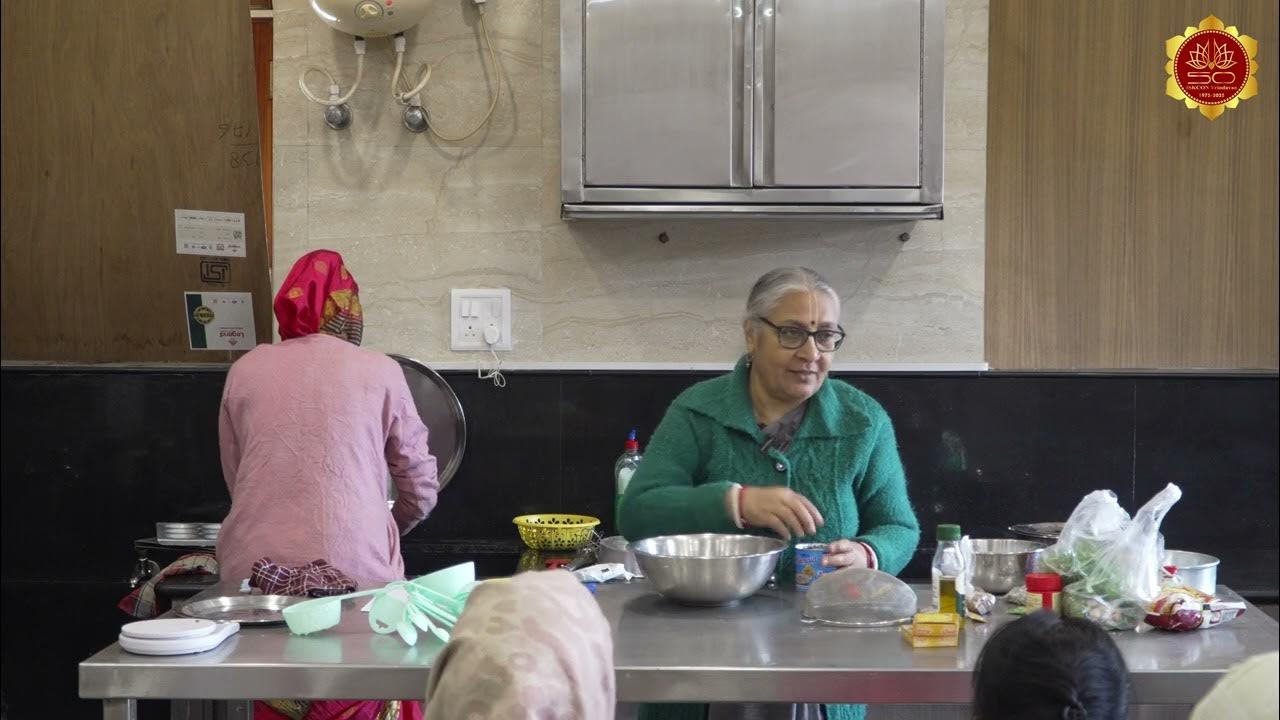 An instructor in a green sweater demonstrates baking at a stainless prep table with bowls, oil, spices, and utensils during a recipe testing session, while a helper works at the stove in a community kitchen.