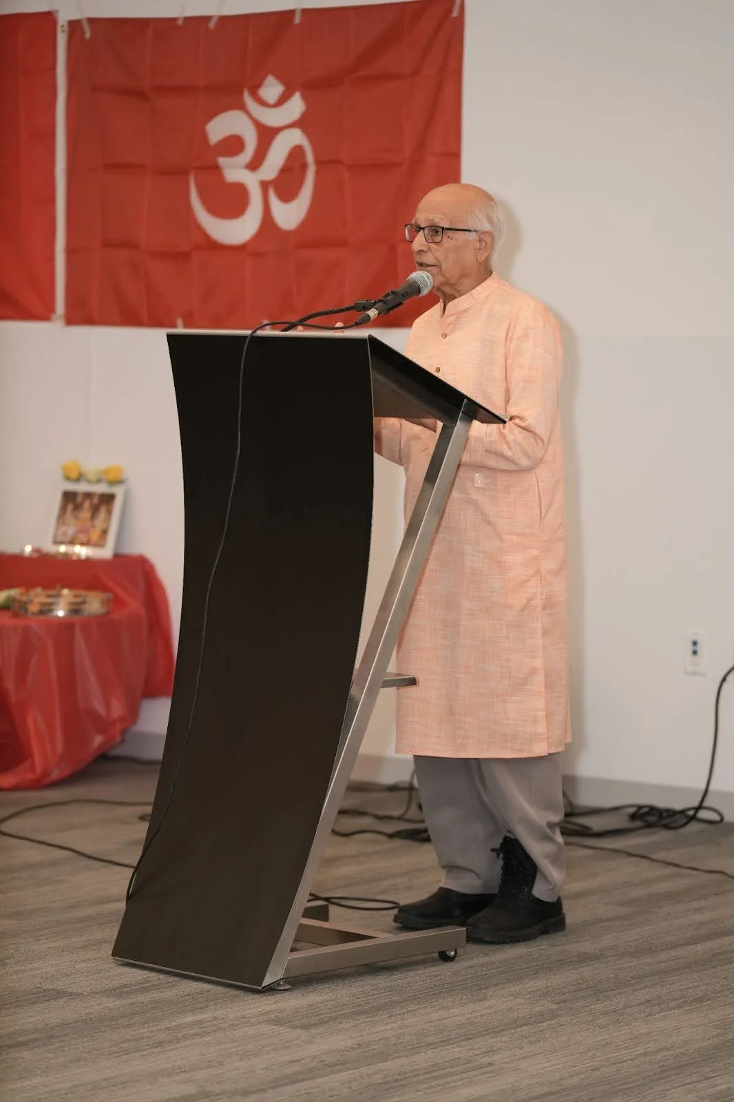 Elderly man in a peach kurta speaks at a podium beside red Om banners and a small puja table, marking Hindu Heritage Month 2025 in Edmonton; community celebration featured on the Cohna Blog.