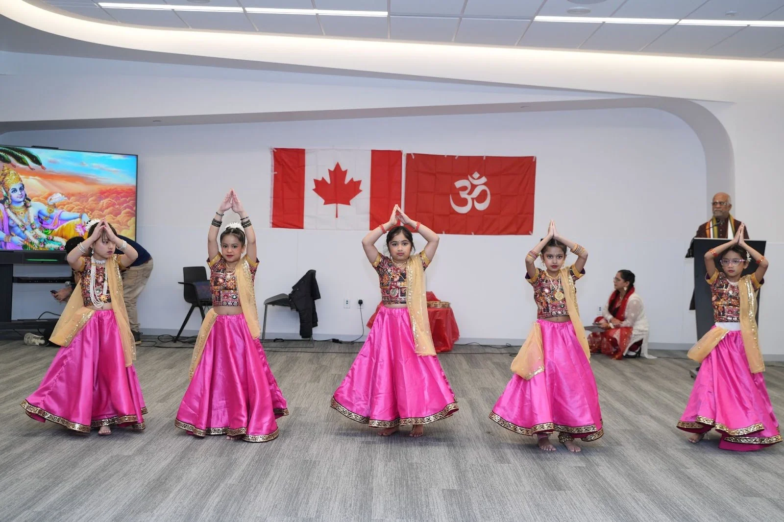 Young girls in bright pink lehengas perform a traditional Indian dance in Edmonton, hands joined in anjali mudra, with Canadian and Om flags behind them during Hindu Heritage Month 2025.