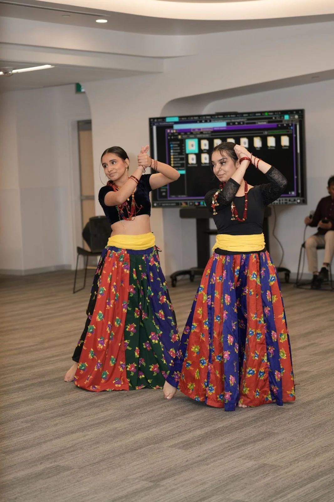 Two dancers perform a traditional Indian folk routine in floral skirts and black tops, arms raised and crossed, at an indoor event in Edmonton during Hindu Heritage Month 2025. Cohna Blog.