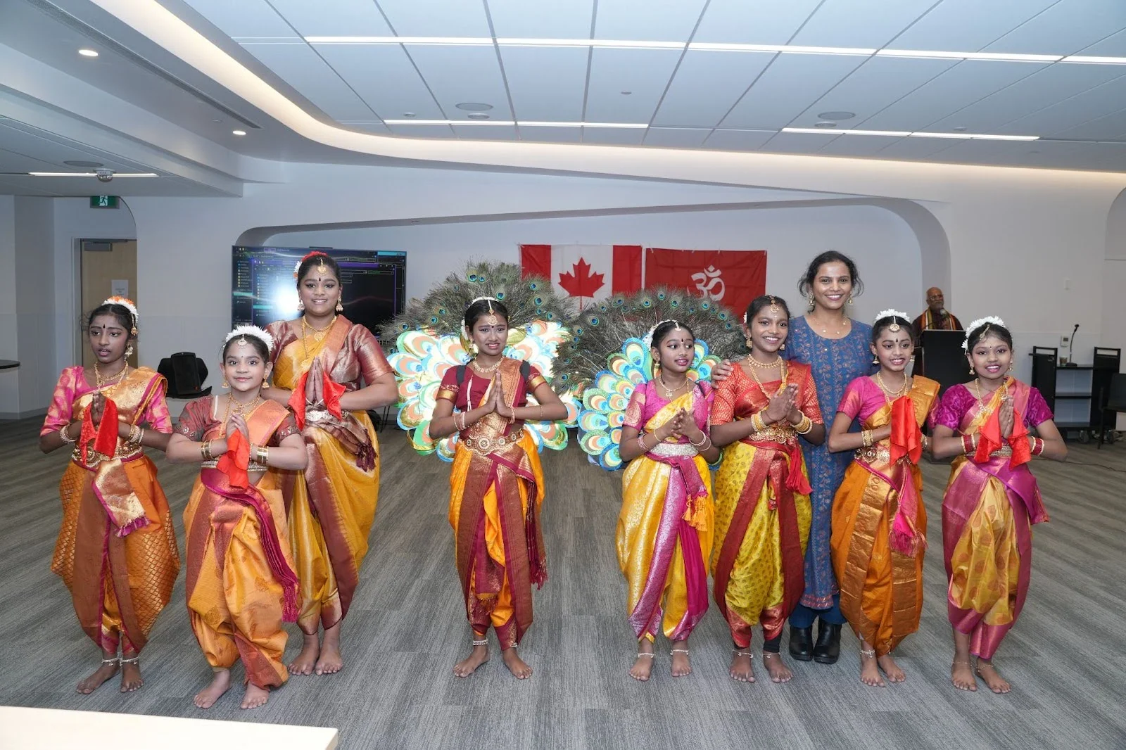 Group of young dancers in vibrant silk costumes pose in namaste at an indoor event in Edmonton, with peacock-feather wings and Canadian and Om flags behind them, celebrating Hindu Heritage Month 2025.