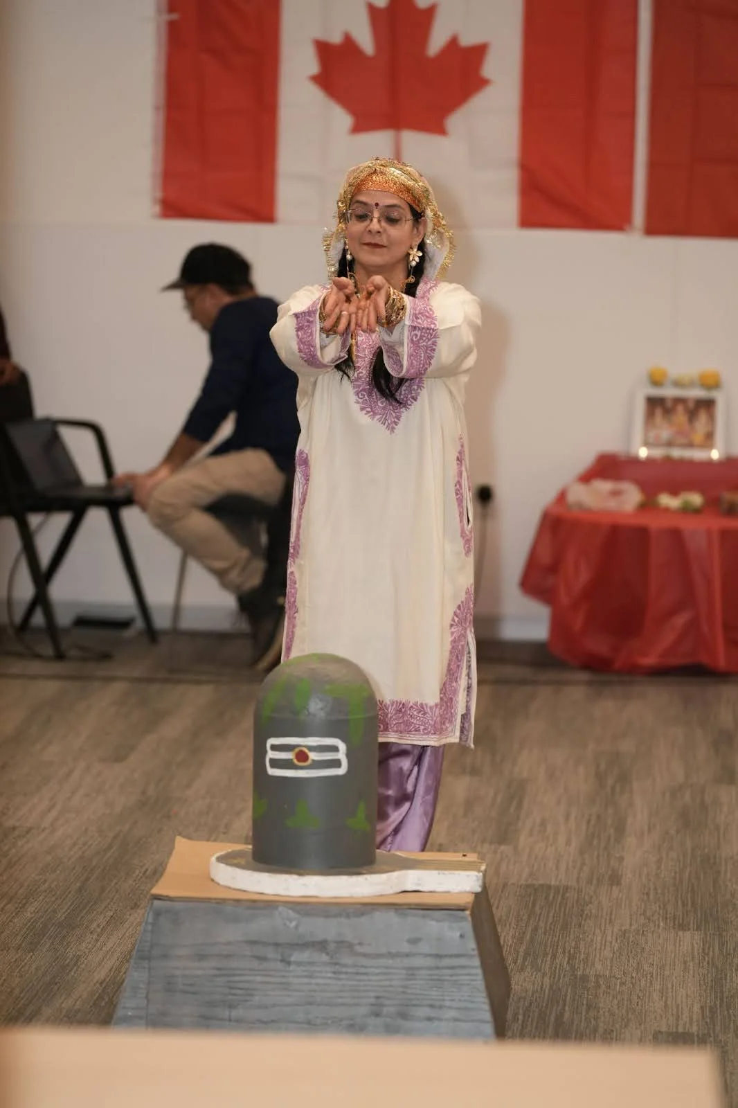 A person in traditional South Asian attire offers a prayer gesture before a grey Shiva Lingam in a community hall, with a large Canadian flag and a small altar visible in the background.