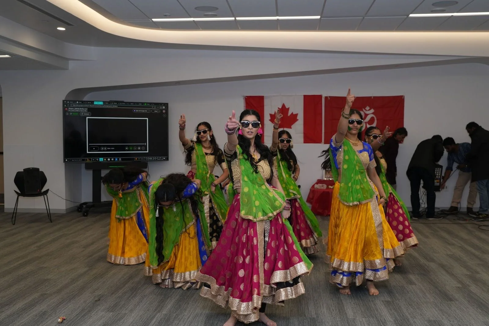 Group of dancers in colorful lehengas and green dupattas perform a traditional Indian dance indoors, with a Canadian flag and red Om flag behind them at a Hindu Heritage Month event in Edmonton.