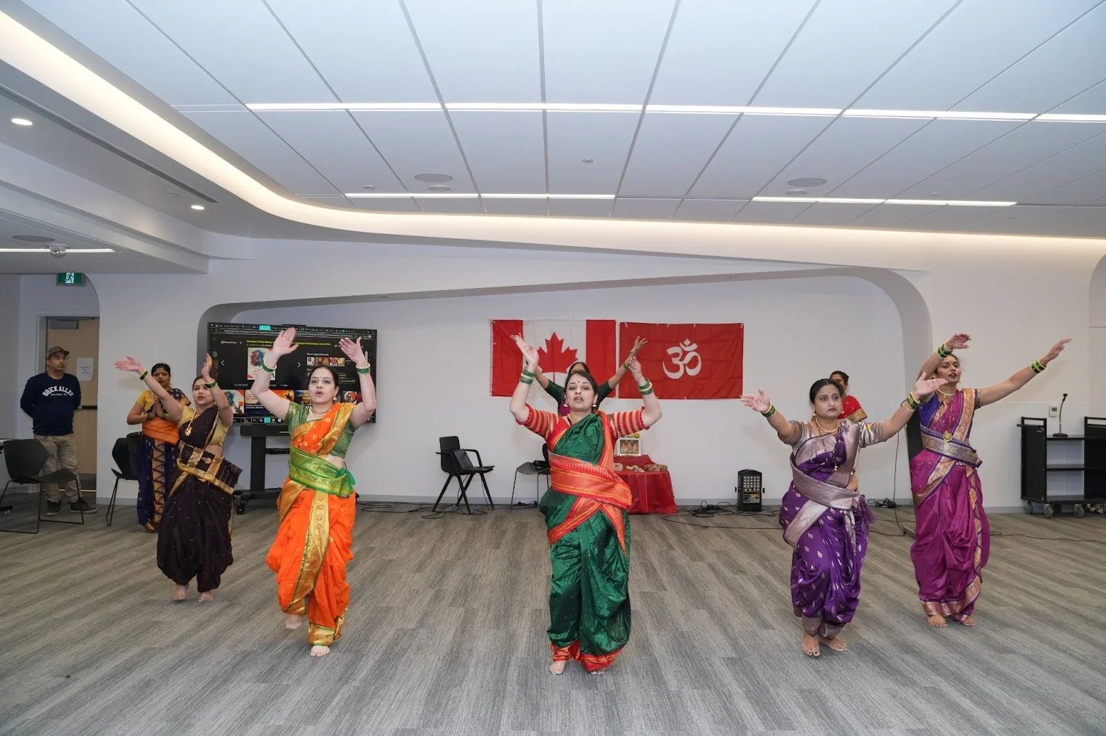 Dancers in vibrant traditional sarees perform a classical routine in a modern hall, arms raised, with Canadian and Om flags and a small devotional table behind them in Edmonton.