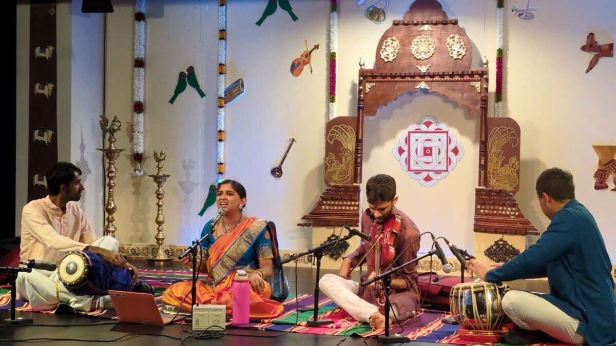 On a temple-style stage, a Carnatic ensemble—sari-clad singer, violinist, and hand-drum percussionists—plays before brass lamps, parrots, and a Sri Yantra, evoking Hindu Marghazi devotion and Living Dharma.