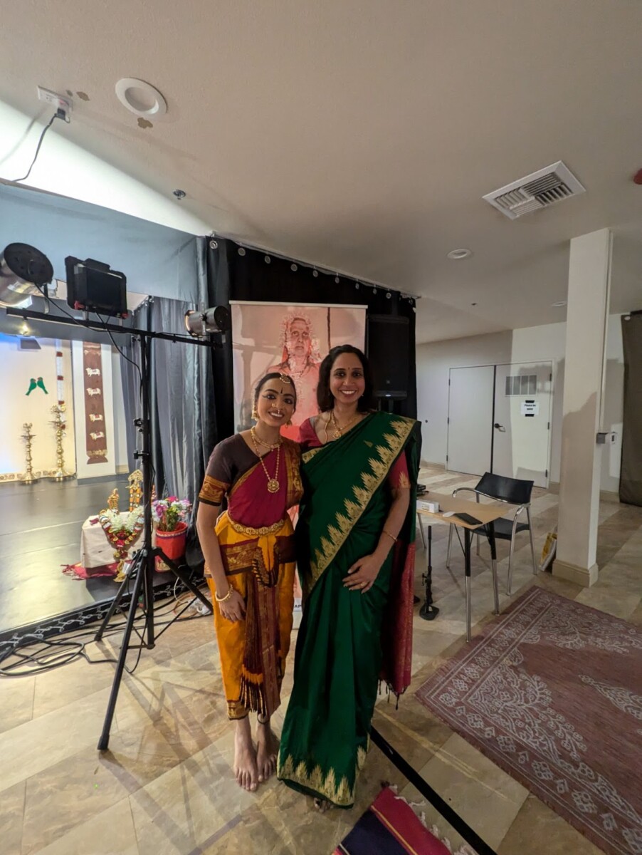 Bharatanatyam dancer in maroon and mustard costume stands barefoot beside a woman in a green sari, near a small stage with lamps, flowers, and a saint’s portrait—Living Dharma in Marghazi.