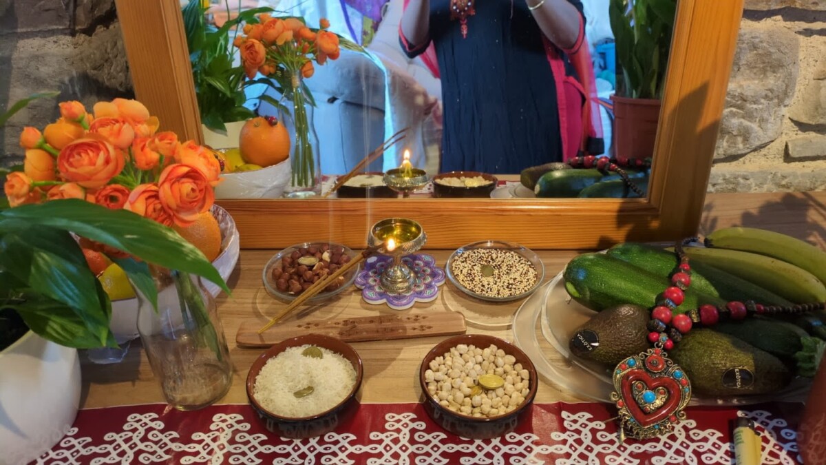 Home puja altar with a lit brass diya before a mirror, bowls of rice, chickpeas, grains, nuts, incense, orange flowers, fruits and vegetables, and a red-beaded mala, evoking Marghazi devotion.