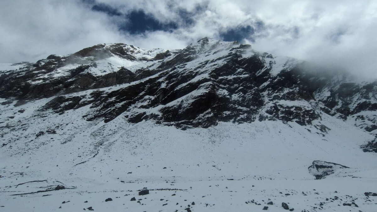 Wide view of a snow-covered mountain with dark rock bands, swirling mist, and overcast clouds; a rocky foreground leads to steep ridges, capturing the stillness and austerity of winter.