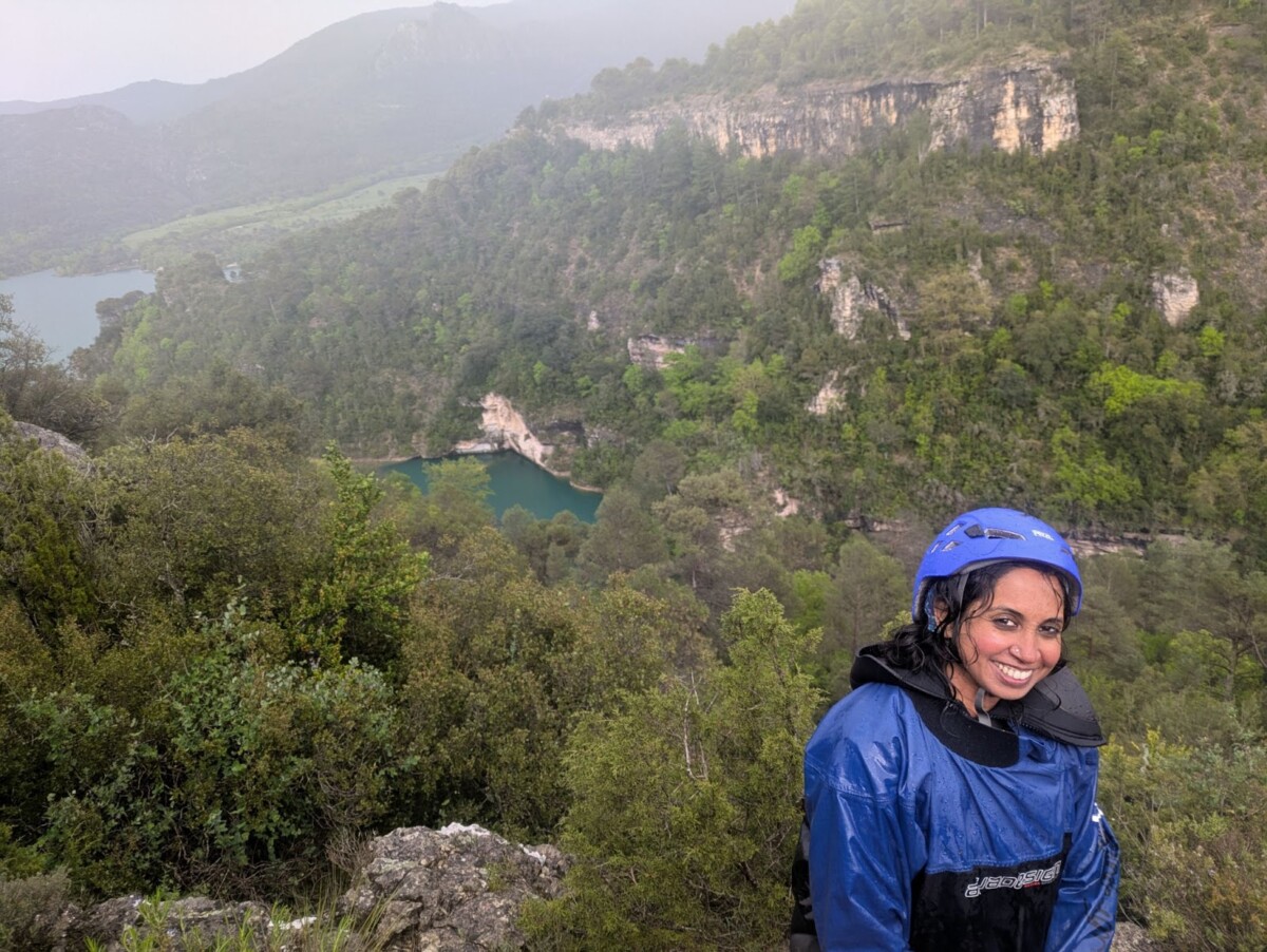 Smiling woman in a blue rain jacket and helmet stands on a rocky overlook after a wet hike, with misty green cliffs, dense pine forest, and a turquoise lake in a deep valley behind her.