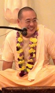 Smiling person in saffron robes with a flower garland sits cross‑legged and speaks into a microphone indoors, captured during a talk on matter and karma for the Articles section.