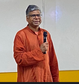 A speaker in an orange kurta holds a handheld microphone before a whiteboard during an indoor seminar, addressing an unseen audience in a classroom setting about culture and media.