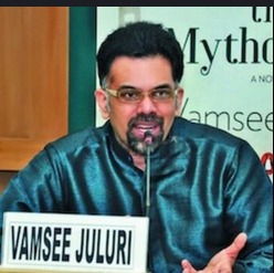 A speaker in a dark silk kurta addresses a tabletop microphone at a book event; a placard reading “VAMSEE JULURI” and a poster with the word “Myth” are visible, indicating a media and culture discussion.