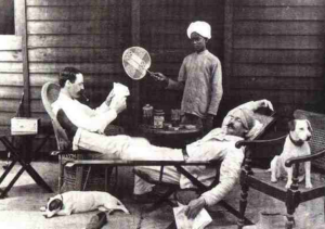 Colonial-era veranda: two men lounge as a turbaned attendant fans them; dogs rest nearby. A stark view of colonial racism and hierarchy tied to narratives about Colonised Indians and Hinduphobia.