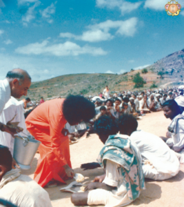 Rural India scene: a saffron-robed spiritual figure with a frizzy hairstyle serves food to seated villagers as volunteers carry pots; a large crowd and blue sky stretch behind.