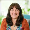 Smiling adult with brown hair in an orange cardigan sits on a teal chair indoors, with plants and soft light; blog cover for a post on friendship anxiety, anxious attachment, and boundaries.