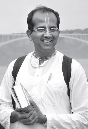 Black-and-white outdoor portrait of a smiling adult holding books, wearing simple traditional clothing with a vertical forehead marking and backpack straps, with a blurred bridge behind; testing keywords.