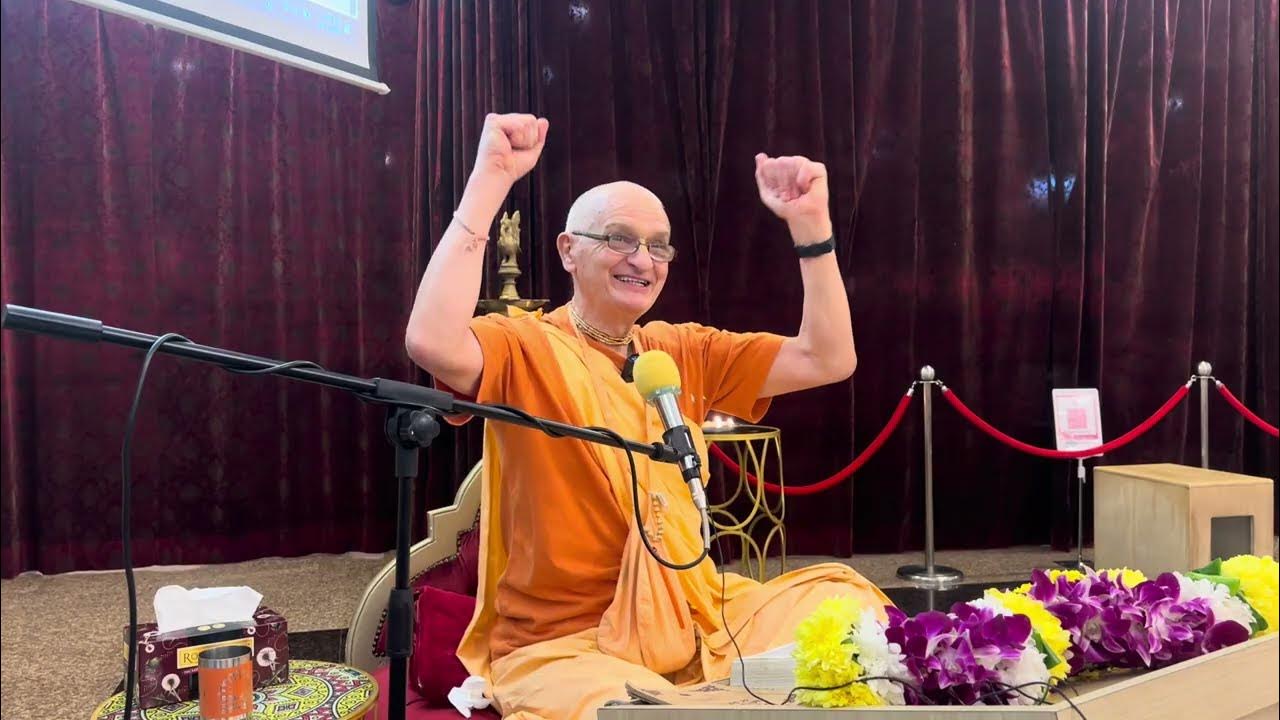 A saffron-robed spiritual speaker smiles and raises both arms during a Bhagavad Gita 2.7 lecture at ISKCON Sri Jagannatha Mandir Kuala Lumpur, seated at a microphone with flowers and a red curtain backdrop.