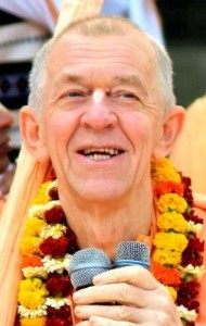 Elderly speaker wearing flower garlands holds two microphones, smiling during a devotional class on the appearance day of Lord Nityananda; close-up portrait, warm colors, event lecture setting.