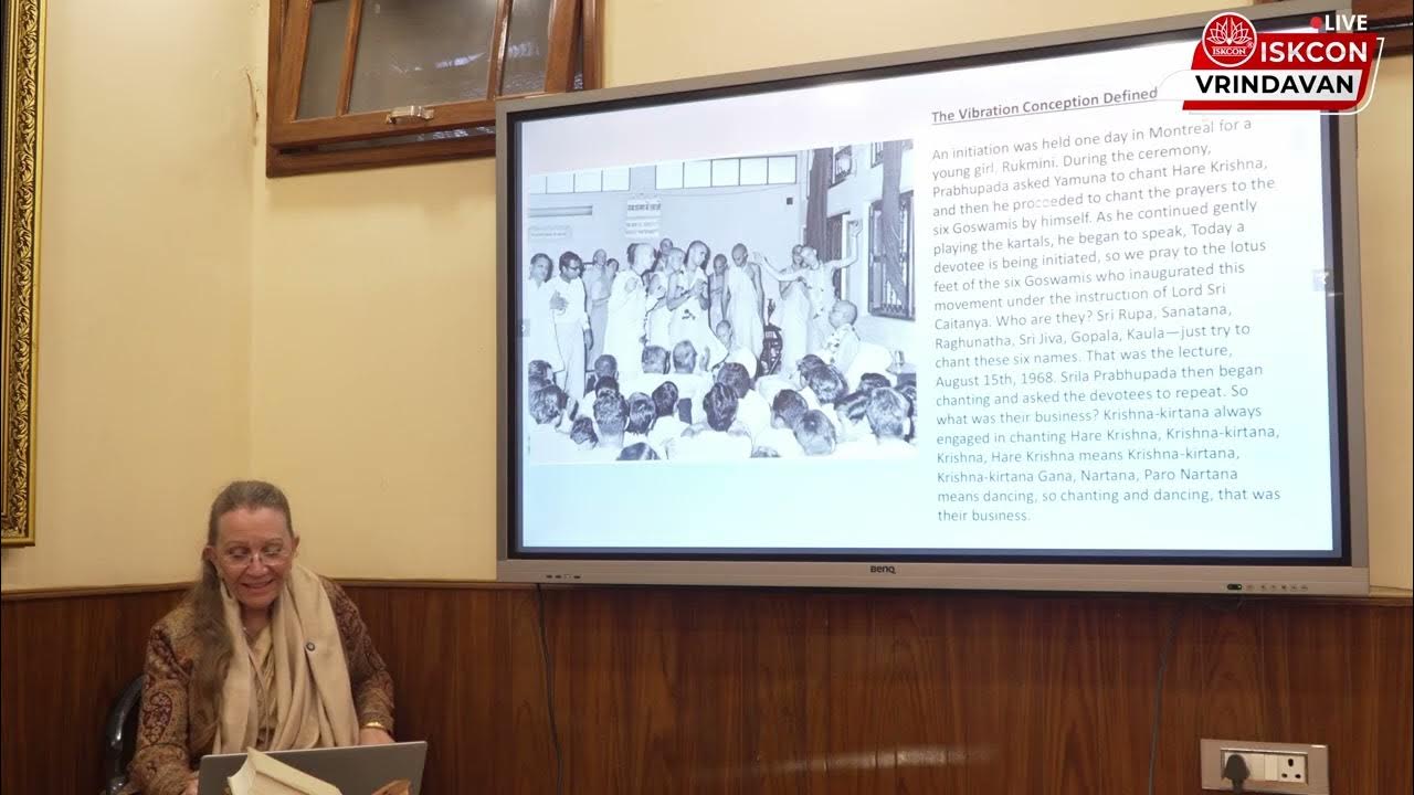 Live ISKCON Vrindavan lecture: a speaker sits beside a large screen showing a slide from Srila Prabhupada Lilamrita with a historic black-and-white gathering and text in a wood-paneled room; educational, testing.