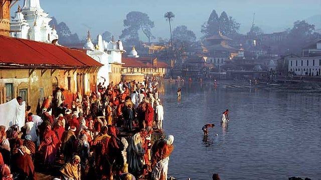 Pilgrims crowd a riverside ghat at dawn, saffron shawls bright against temple spires and misty hills; some bathe in calm water during rituals—an evocative scene of Bharatavarsha’s sacred bonds and pilgrimage.