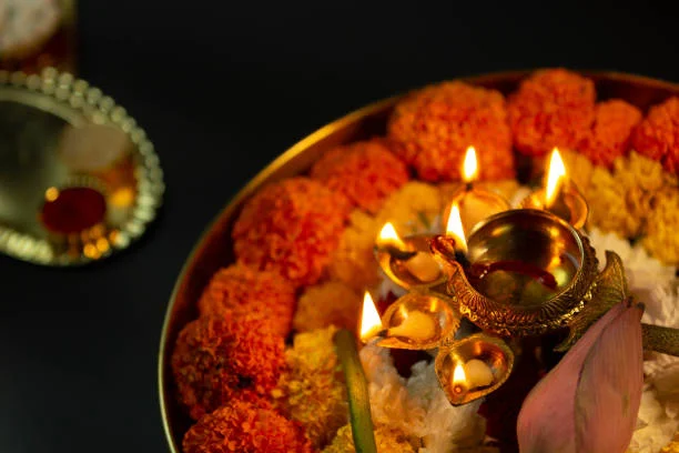 Lit brass multi-wick diya encircled by orange and yellow marigolds and white jasmine on a puja thali, with a lotus bud and kumkum tray, symbolizing Hindu New Year rituals and Festivals celebration.